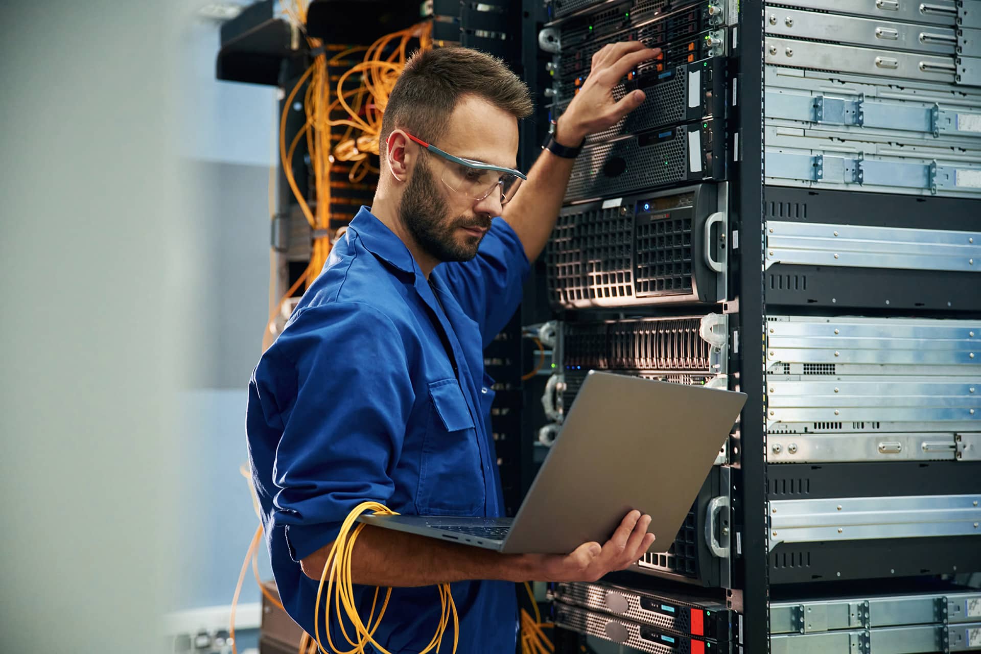 Silver colored laptop in hands. Young man is working with internet equipment and wires in server room.
