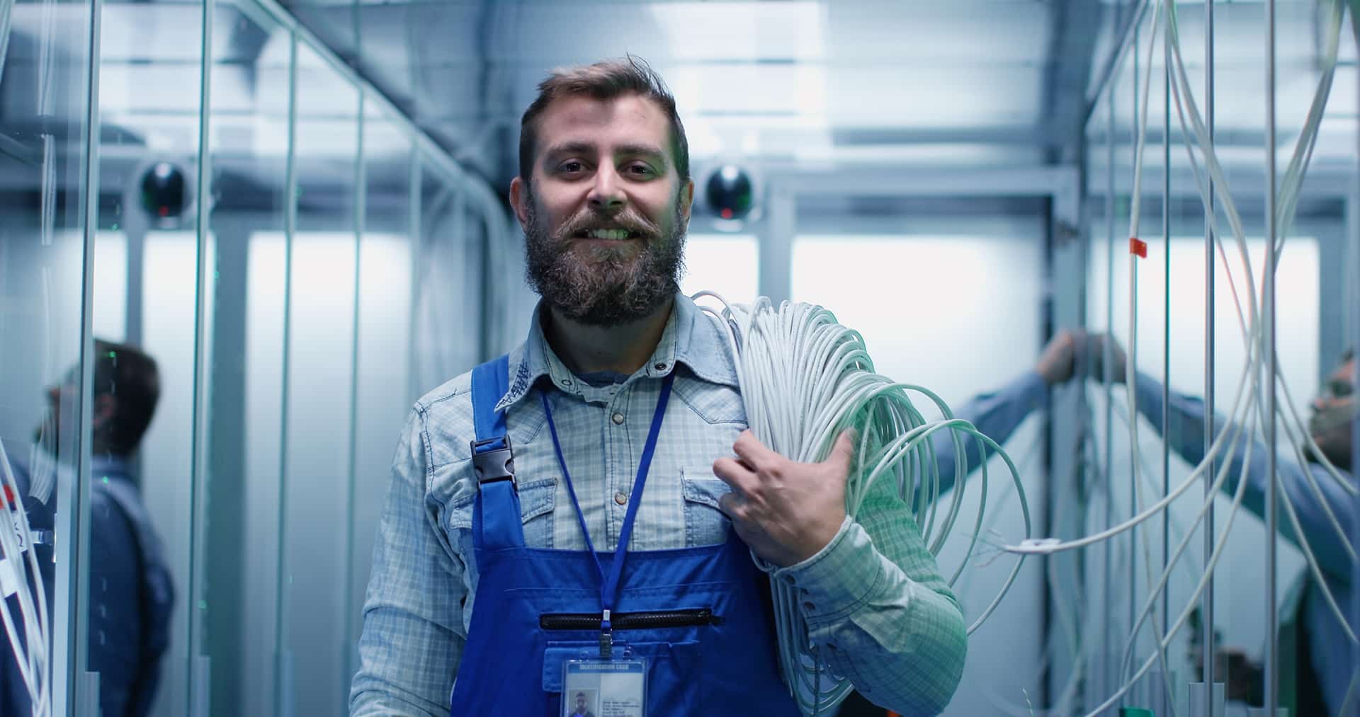 Portrait of adult bearded man technician standing in corridor with cable on his shoulders of server room in data center smiling at camera